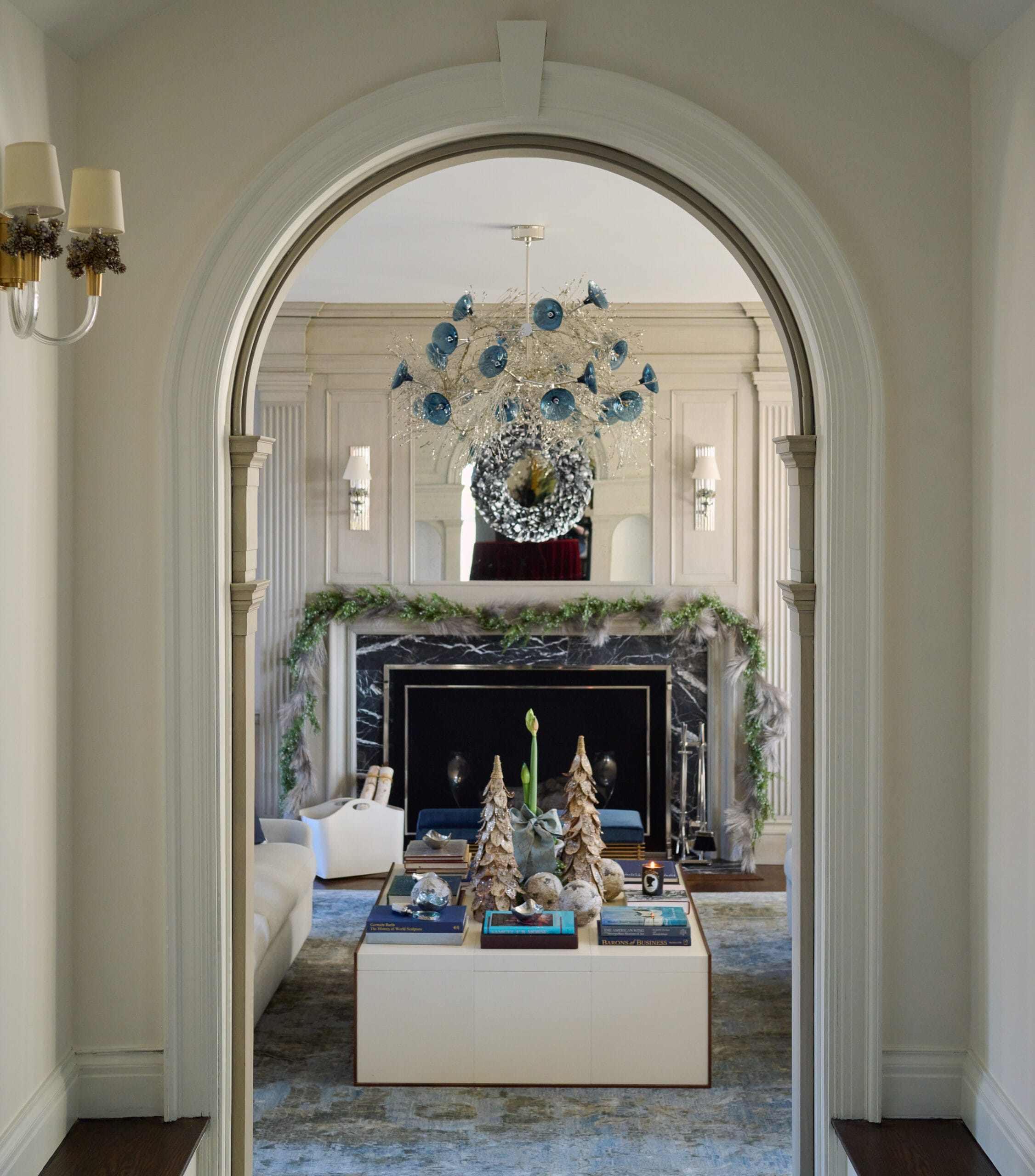Living room decorated with holiday decor in a soft blue color palette. Wreath over the mantel. Christmas tree decorations sit on the coffee table for a warm festive environment.