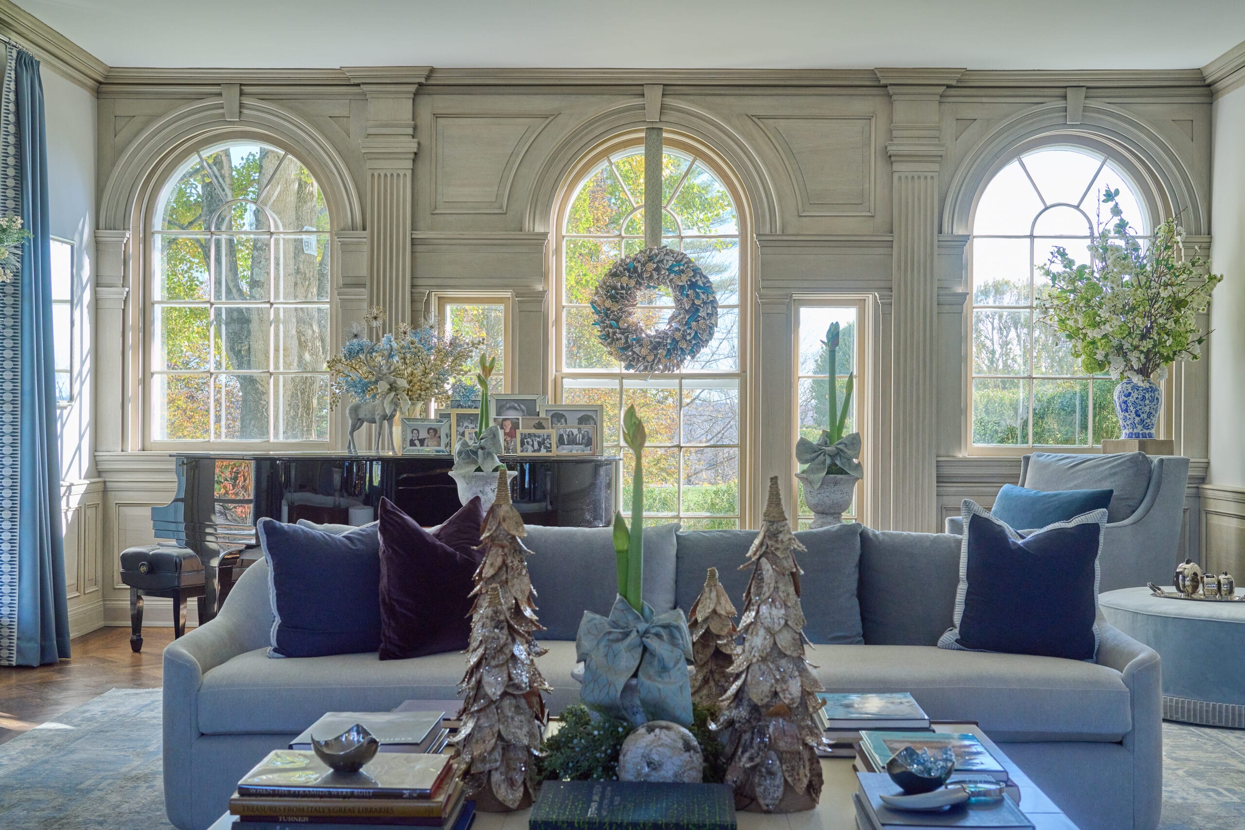 Living room decorated with holiday decor in a soft blue color palette. A wreath hangs over the window. Four christmas tree decorations sit on the coffee table.